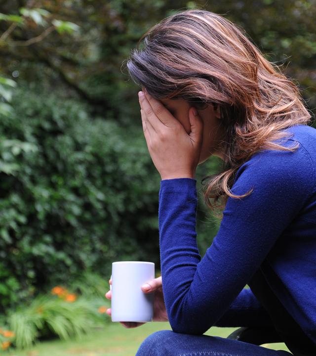Frau sitzt mit Tasse draußen und hält sich mit einer Hand den kopf. Sie scheint deprimiert zu sein oder Schmerzen zu haben.
