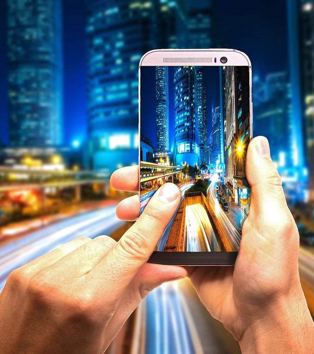 Nächtlicher Straßenzug, im Vordergrund eine Hand mit Handy
