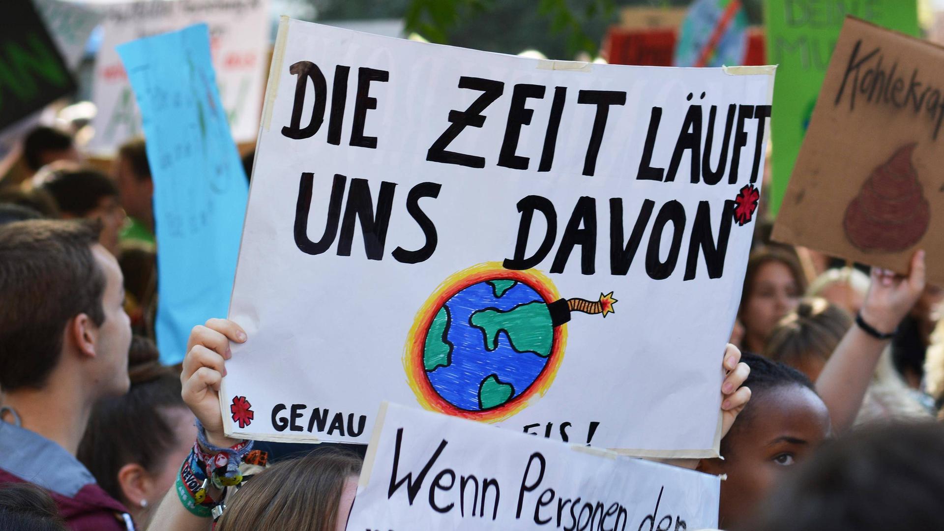 Viele Demonstrierende halten Plakate für mehr Klimaschutz in die Luft. Im Mittelpunkt erkennt man ein großes Schild, mit der Aufschrift "Die Zeit läuft uns davon". Außerdem ist eine explodierende und brennen Weltkugel auf das Plakat gemalt.