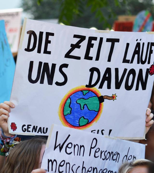 Viele Demonstrierende halten Plakate für mehr Klimaschutz in die Luft. Im Mittelpunkt erkennt man ein großes Schild, mit der Aufschrift "Die Zeit läuft uns davon". Außerdem ist eine explodierende und brennen Weltkugel auf das Plakat gemalt.