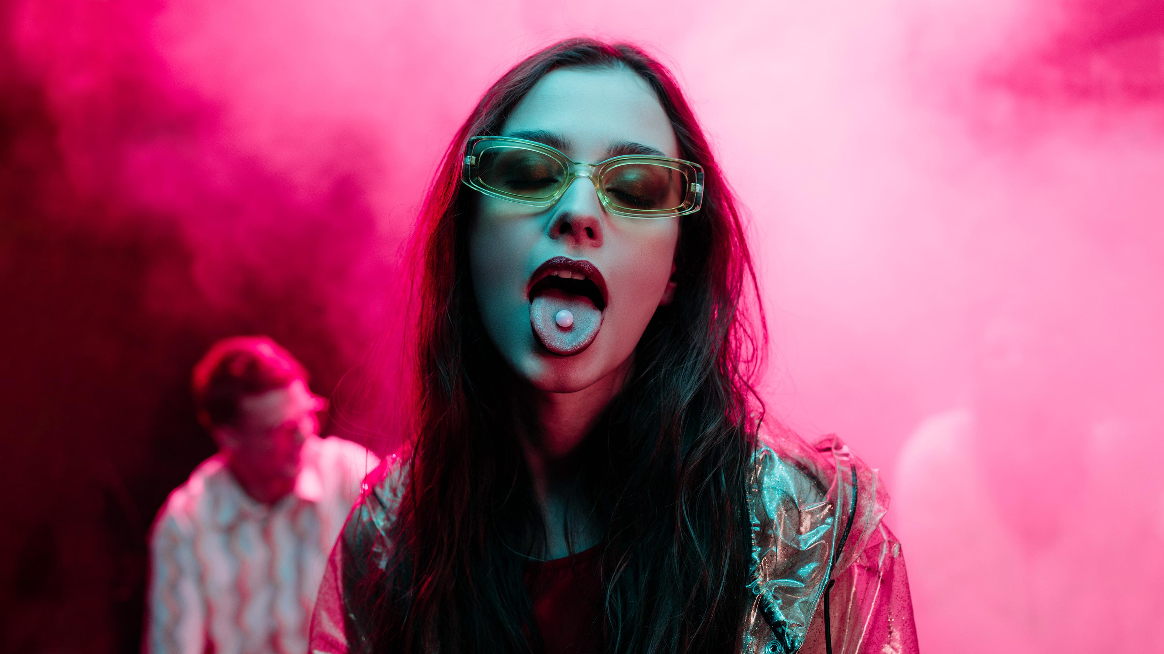 Frau mit langen Haaren und Brille vor pinkem Hintergrund streckt die Zunge raus, auf der eine kleine Pille liegt.