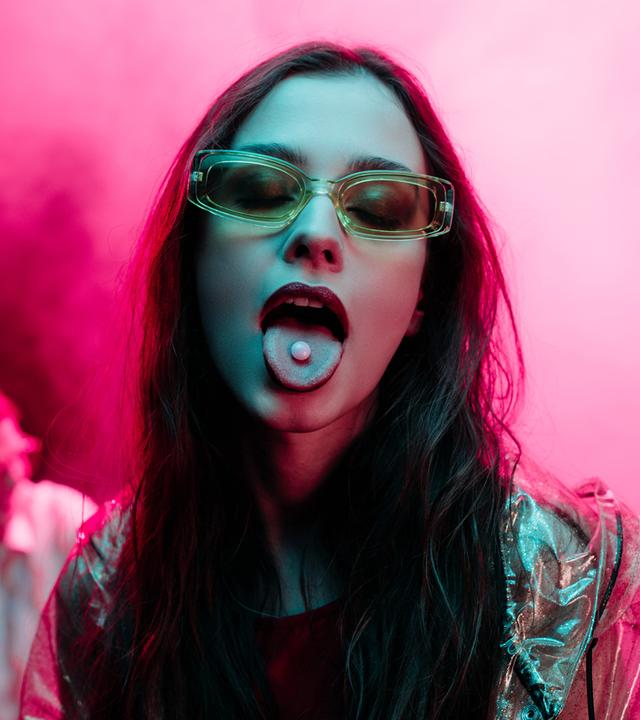 Frau mit langen Haaren und Brille vor pinkem Hintergrund streckt die Zunge raus, auf der eine kleine Pille liegt.