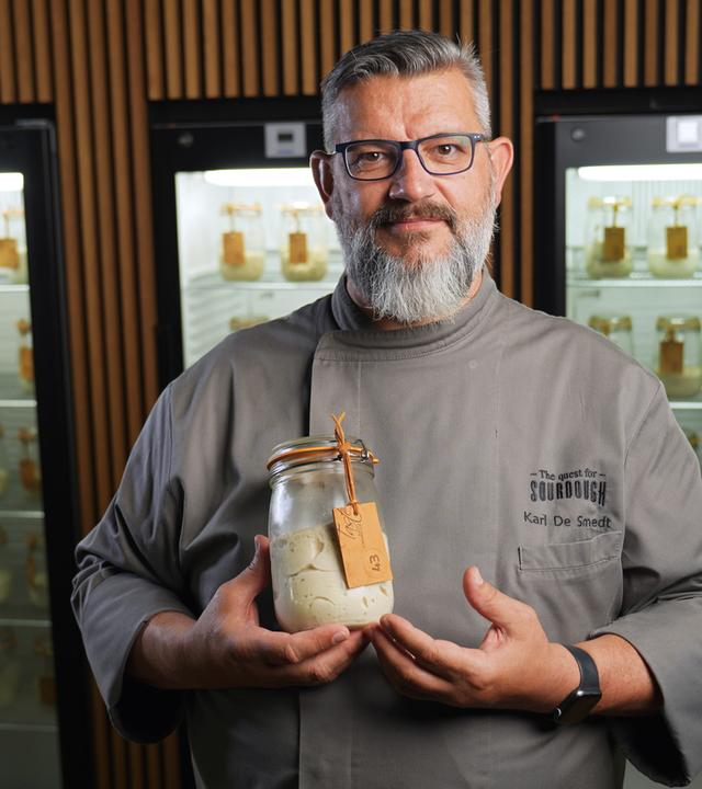 Ein Mann in grauer Kochjacke hält ein Einmachglas mit Sauerteig in der Hand.