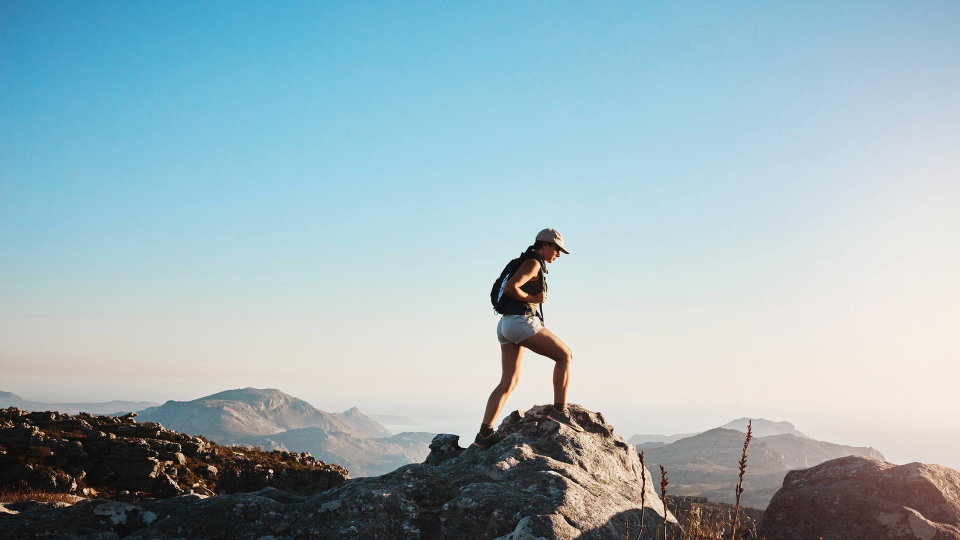 Vor blauem Himmel steht eine Frau auf einer Bergspitze und schaut ins Tal.