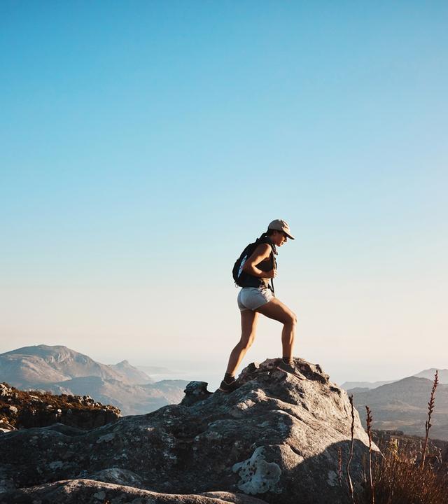 Vor blauem Himmel steht eine Frau auf einer Bergspitze und schaut ins Tal.
