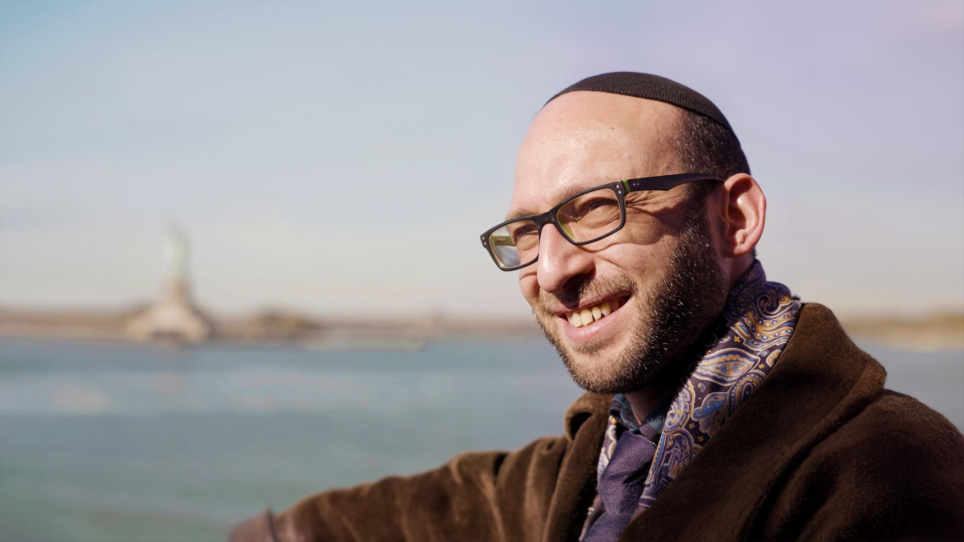 Rabbi Akiva Weingarten (39) lächelt im Vordergrund, während im Hintergrund die Freiheitsstatue und der Hudson River in New York unscharf zu sehen sind.