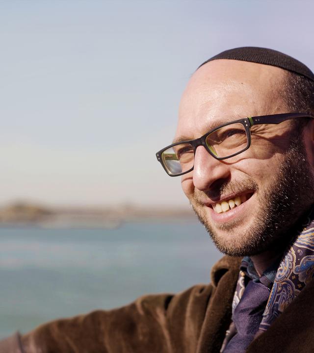 Rabbi Akiva Weingarten (39) lächelt im Vordergrund, während im Hintergrund die Freiheitsstatue und der Hudson River in New York unscharf zu sehen sind.