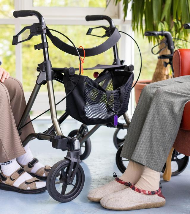 Alte Menschen sitzen auf Sesseln, dazwischen ein  Rollator