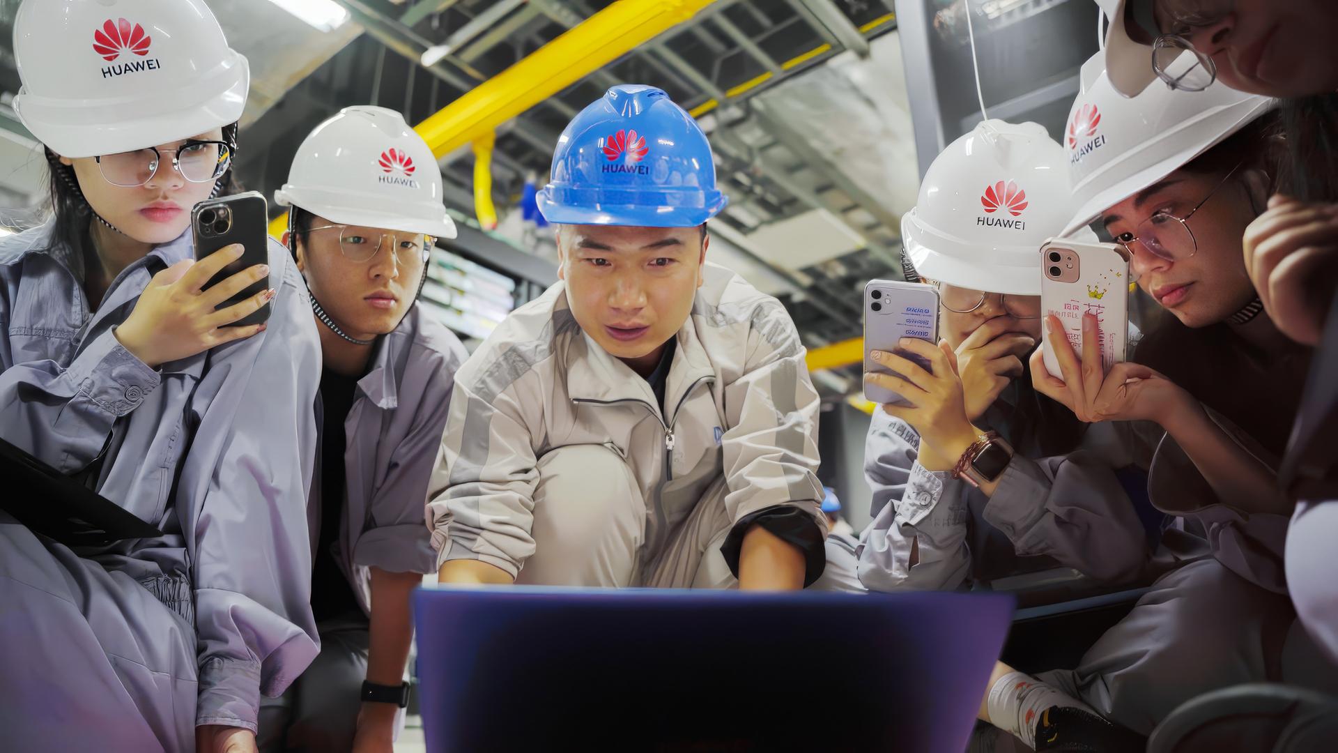 Vier junge Leute mit weißen Helmen mit Huawei-Logo und in hellgrauen Arbeitsoveralls sitzen oder hocken auf dem Boden vor einem Lap-Top, das von einem jungen Mann in mit blauem Helm und weißem Overall ihrer Mitte bedient wird. Drei von ihnen haben ihre Handys filmend auf den Lap-Top-Monitor gerichtet.