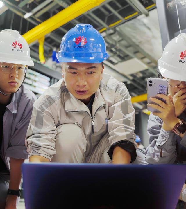 Vier junge Leute mit weißen Helmen mit Huawei-Logo und in hellgrauen Arbeitsoveralls sitzen oder hocken auf dem Boden vor einem Lap-Top, das von einem jungen Mann in mit blauem Helm und weißem Overall ihrer Mitte bedient wird. Drei von ihnen haben ihre Handys filmend auf den Lap-Top-Monitor gerichtet.