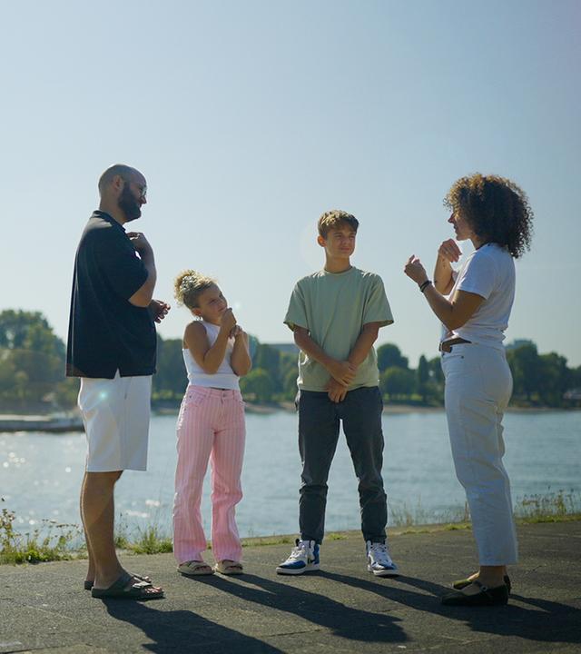 Familie Rose: Eltern mit Behinderung": Familie Rose, die Eltern mit zwei Kindern, steht am Flussufer, Vater und Tochter gebärden.