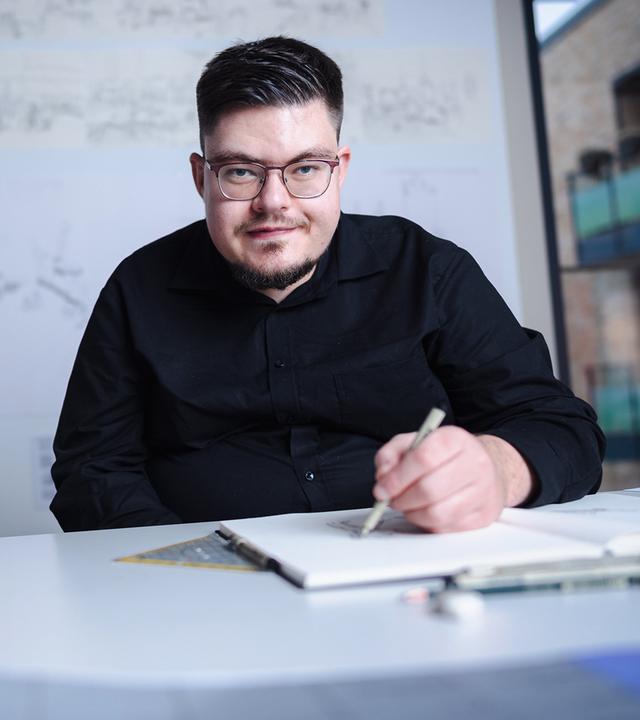 Elias von Martial sitzt an seinem Schreibtisch im Kunsthaus kaethe:k.  Er trägt ein schwarzes Hemd und blickt in die Kamera. 