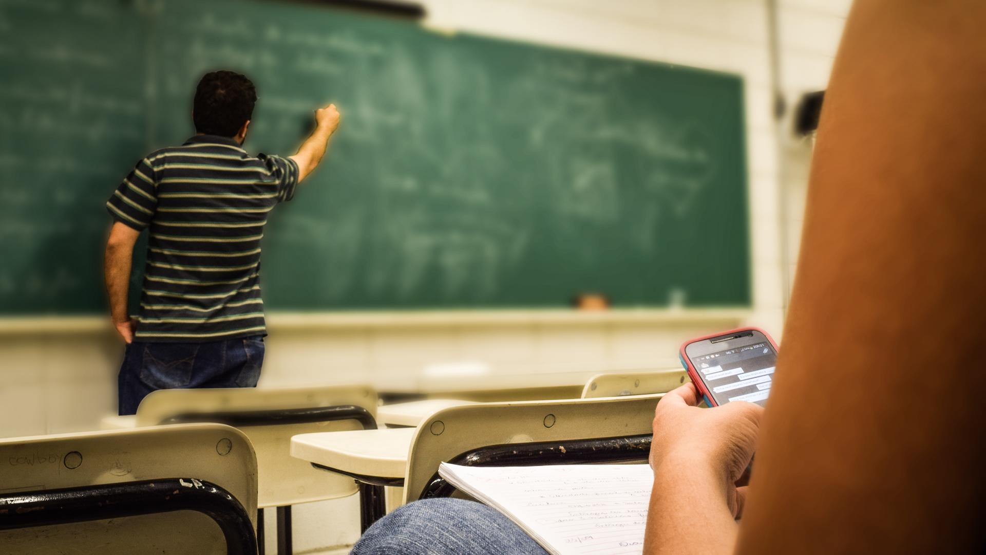 Ein Lehrer erklärt an der Tafel, während ein Schüler neben ihm ein Smartphone nutzt – Symbolbild für den digitalen Wandel und die Herausforderung, KI-Technologie sinnvoll im Unterricht einzusetzen.