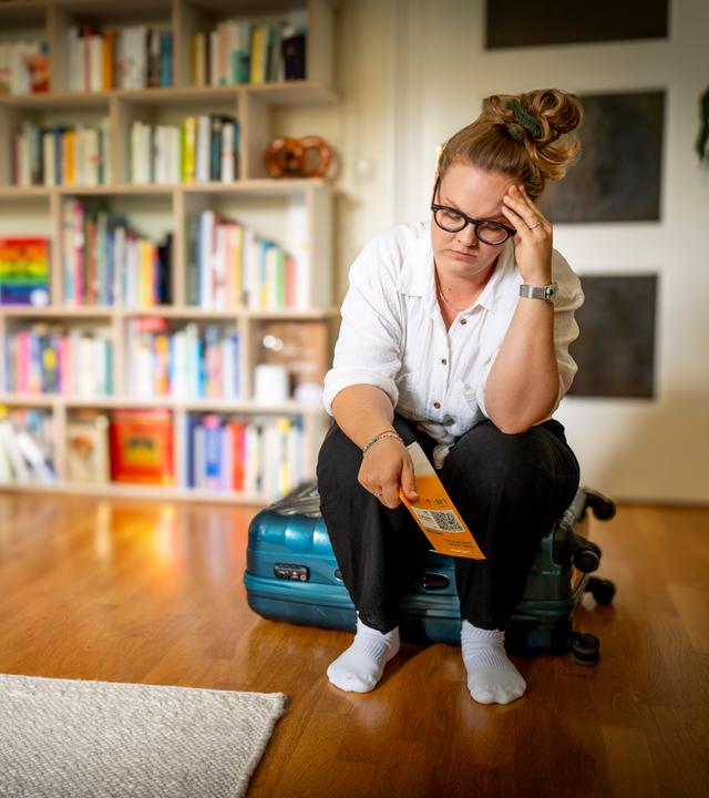 "37°Leben: Wenn die Panik mitfliegt - Flugangst besiegen": Sophie (33) sitzt auf einem Koffer in ihrem Wohnzimmer und hat ein Flugticket in der Hand. Ihr Kopf ist mit einer Hand aufgestützt und ihr Gesichtsausdruck zeigt ihre Sorge und Angst.