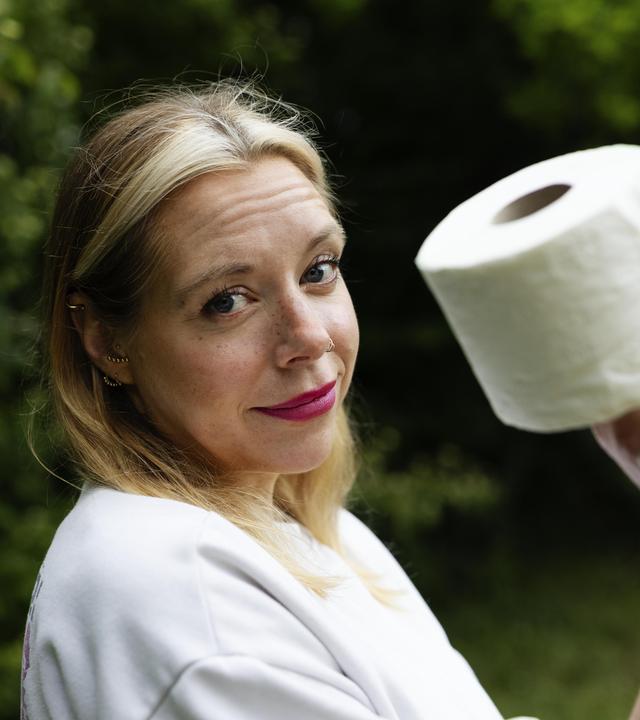 37°Leben: Müssen müssen - jung und inkontinent": Birgit, eine 40-jährige Frau mit langen blonden Haaren und einem Nasenpiercing, steht seitlich zur Kamera vor grünen Büschen. In der rechten Hand hält sie eine Rolle Toilettenpapier. Ihr Blick ist skeptisch bis fragend, dabei schmunzelt sie ein bisschen.