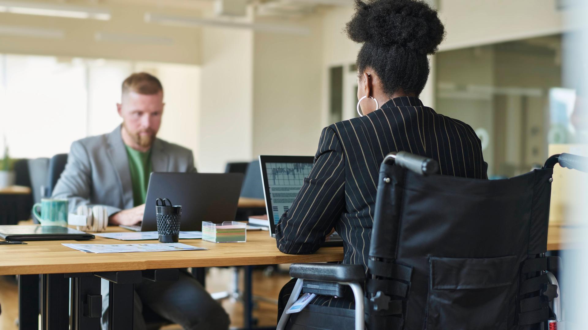 Ein weißer Mann und eine schwarze Kollegin im Rollstuhl sitzen sich an einem Schreibtisch im Büro gegenüber.
