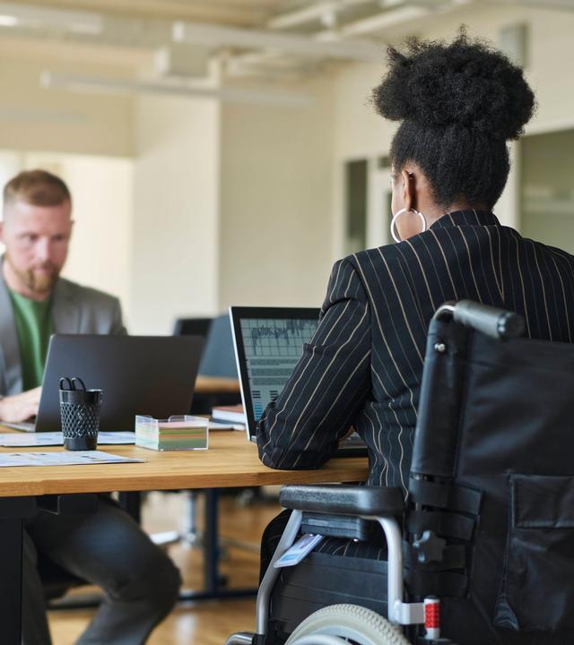 Ein weißer Mann und eine schwarze Kollegin im Rollstuhl sitzen sich an einem Schreibtisch im Büro gegenüber.