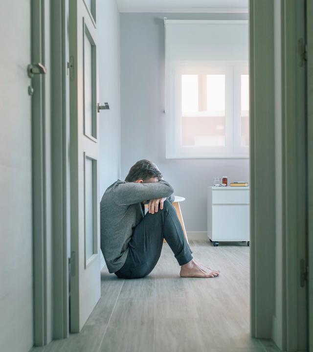 Mann sitzt an die Wand gelehnt auf dem Boden und hält sich die Hände vors Gesicht. 