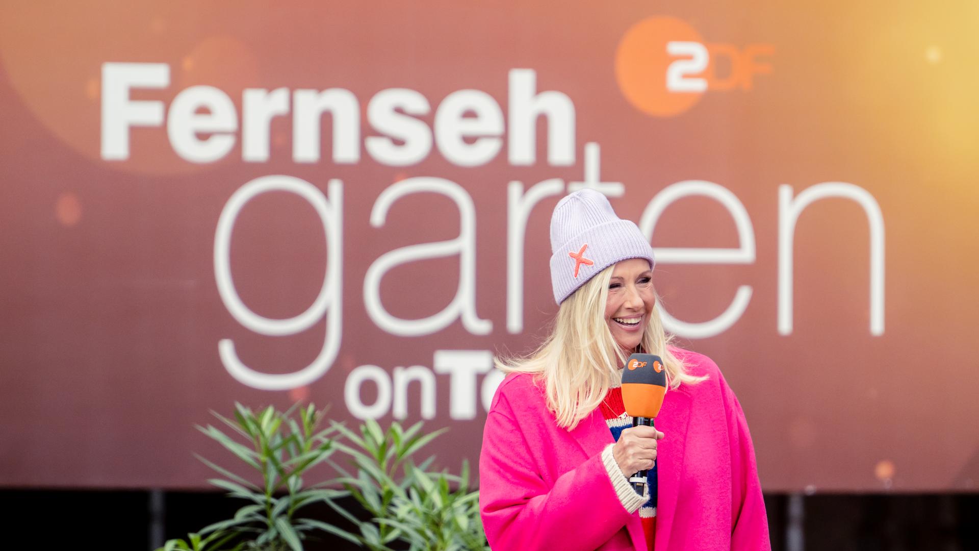 Andrea Kiewel steht vor einer Video-Leinwand, auf der das "ZDF Fernsehgarten"-Logo zu sehen ist.