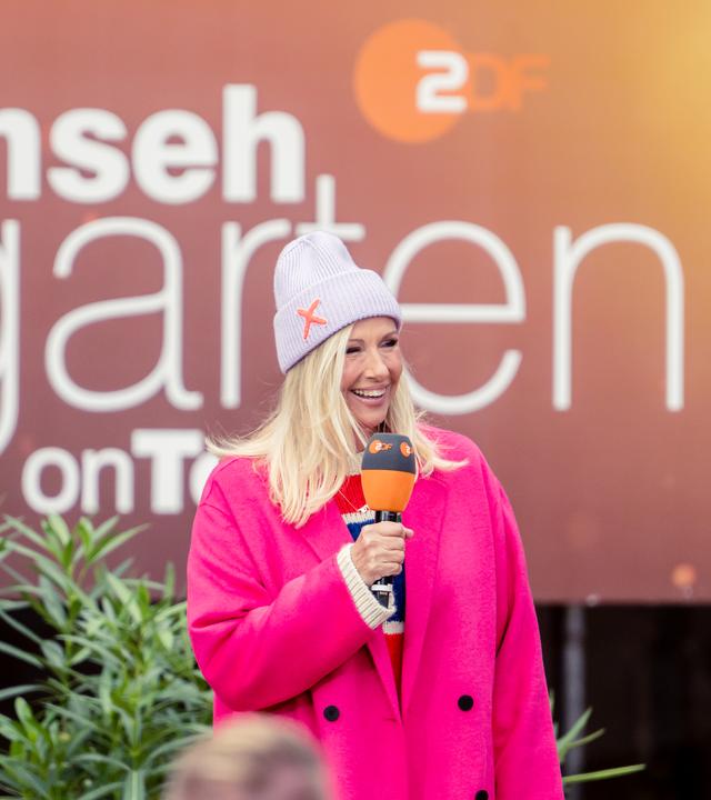 Andrea Kiewel steht vor einer Video-Leinwand, auf der das "ZDF Fernsehgarten"-Logo zu sehen ist.