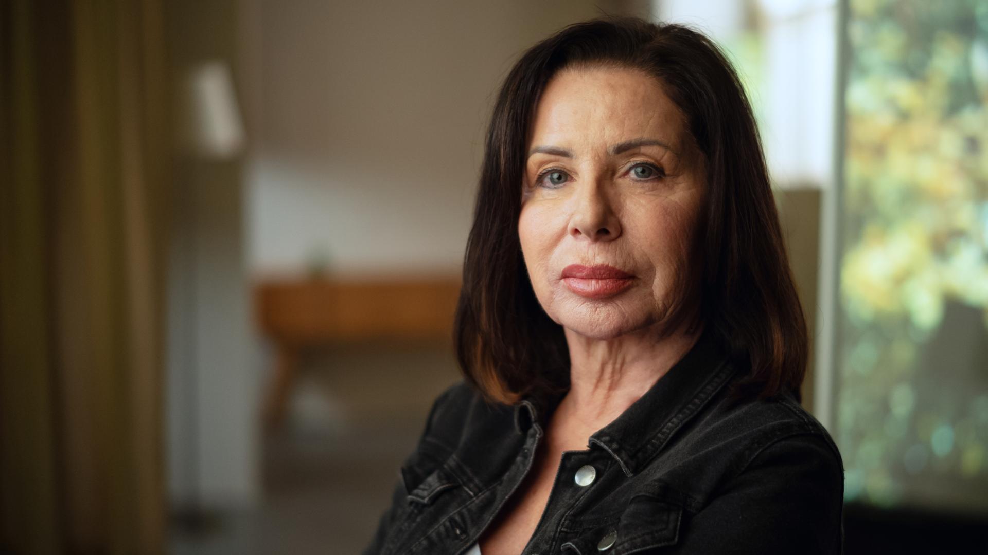 Manuela Freitag im Portrait fotografiert. Sie sitzt mit dem Gesicht der Kamera zugewandt und schaut ernst. Manuela Freitag ist 64 Jahre alt und trägt ihre dunkelbraunen Haare schulterlang. 