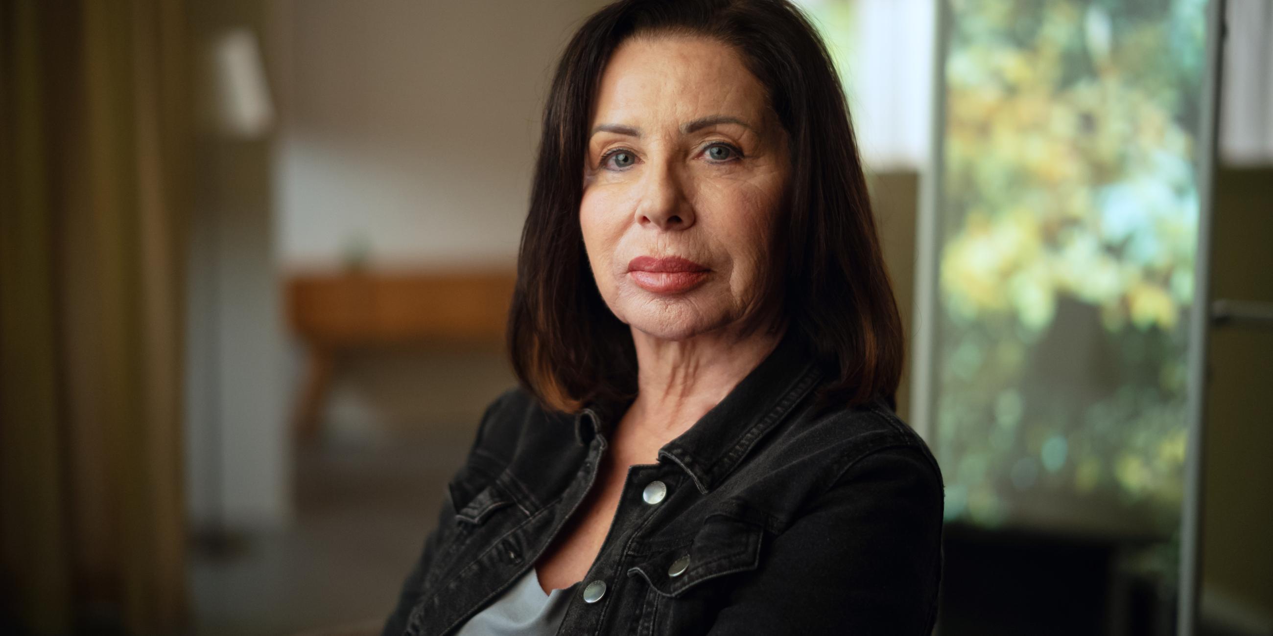 Manuela Freitag im Portrait fotografiert. Sie sitzt mit dem Gesicht der Kamera zugewandt und schaut ernst. Manuela Freitag ist 64 Jahre alt und trägt ihre dunkelbraunen Haare schulterlang. 
