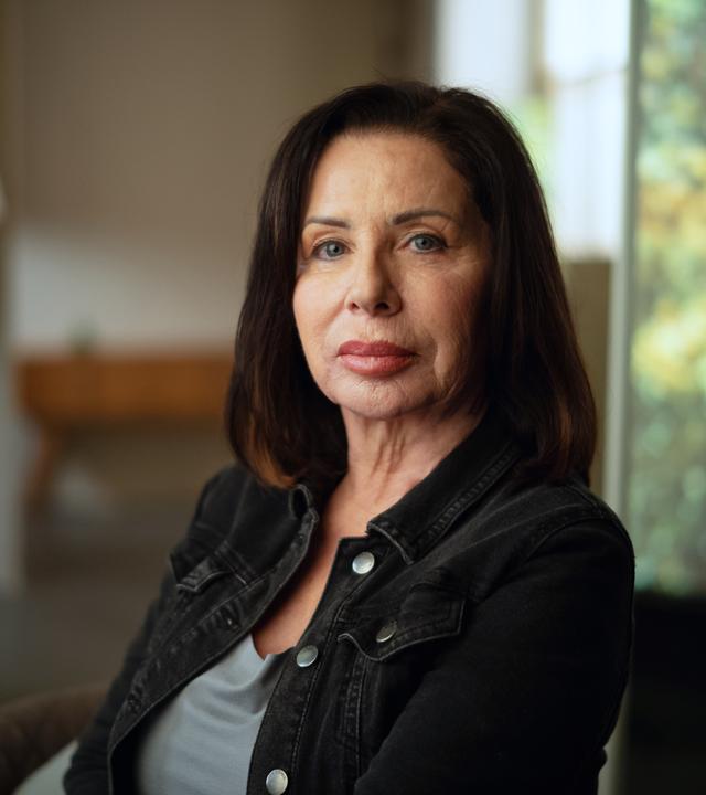 Manuela Freitag im Portrait fotografiert. Sie sitzt mit dem Gesicht der Kamera zugewandt und schaut ernst. Manuela Freitag ist 64 Jahre alt und trägt ihre dunkelbraunen Haare schulterlang. 