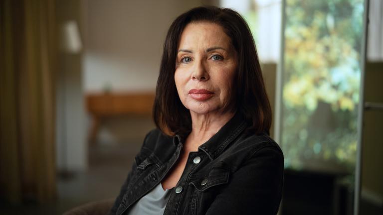 Manuela Freitag im Portrait fotografiert. Sie sitzt mit dem Gesicht der Kamera zugewandt und schaut ernst. Manuela Freitag ist 64 Jahre alt und trägt ihre dunkelbraunen Haare schulterlang. 