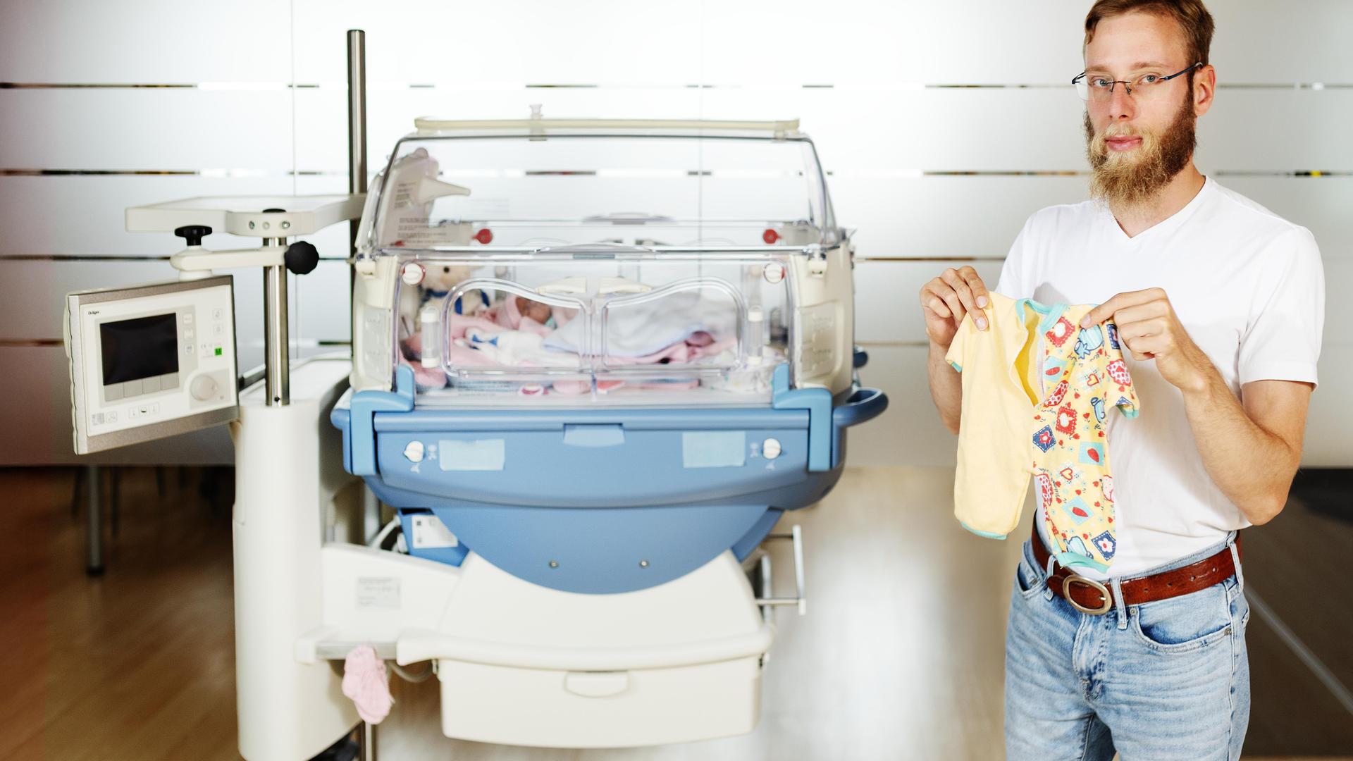 Lukas Mader steht neben einem Brutkasten und hält seinen Strampler hoch. Er selbst ist ein Extremfrühchen - drei Monate zu früh auf die Welt gekommen.