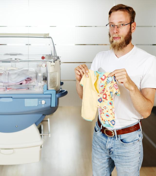 Lukas Mader steht neben einem Brutkasten und hält seinen Strampler hoch. Er selbst ist ein Extremfrühchen - drei Monate zu früh auf die Welt gekommen.