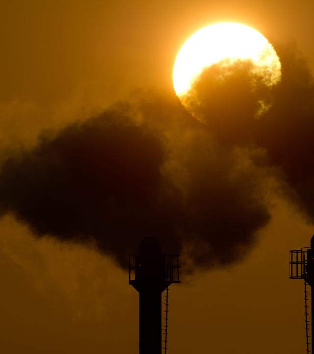  Firmen zahlen für CO2-Emissionen und sparen bei weniger Ausstoß. Auf dem Bild hält ein Mann ein CO2-Schild, während eine Autoschlange Abgase ausstößt.