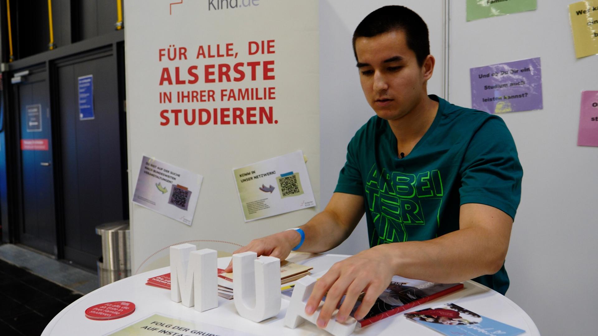  Christopher Khlongklad (23) trägt ein dunkelgrünes T-Shirt von Arbeiterkind.de und steht am Messestand von Arbeiterkind.de. Dabei richtet er den Buchstaben "T" auf dem Tisch auf, der das aus weißen Buchstaben bestehende Wort "Mut" vollendet. 