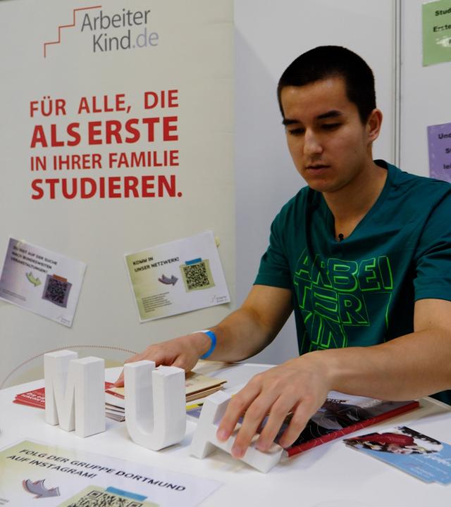  Christopher Khlongklad (23) trägt ein dunkelgrünes T-Shirt von Arbeiterkind.de und steht am Messestand von Arbeiterkind.de. Dabei richtet er den Buchstaben "T" auf dem Tisch auf, der das aus weißen Buchstaben bestehende Wort "Mut" vollendet. 