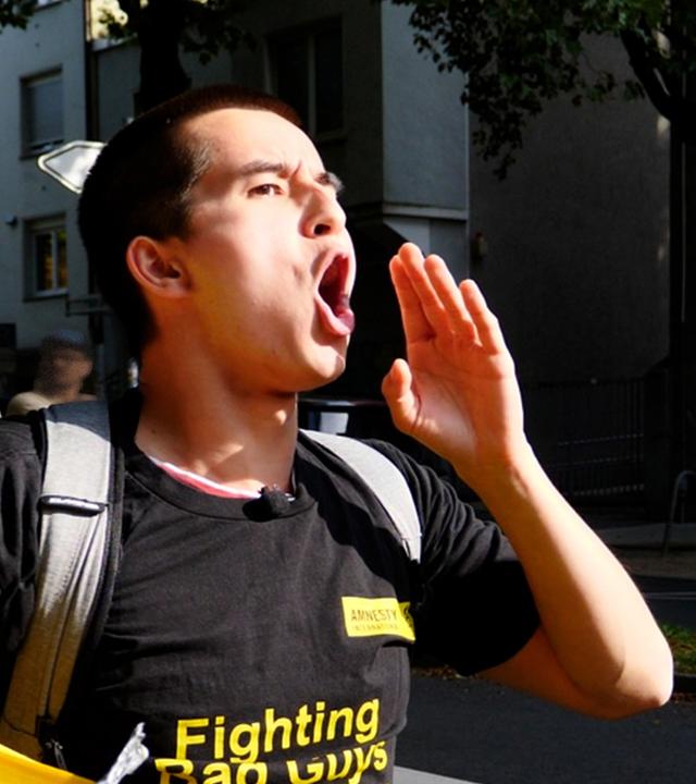 Christopher (23) trägt ein schwarzes T-Shirt und hält mit der rechten Hand ein gelbes Amnesty- Banner. Er hat die linke Hand an den Mund gelegt und ruft laut mit.