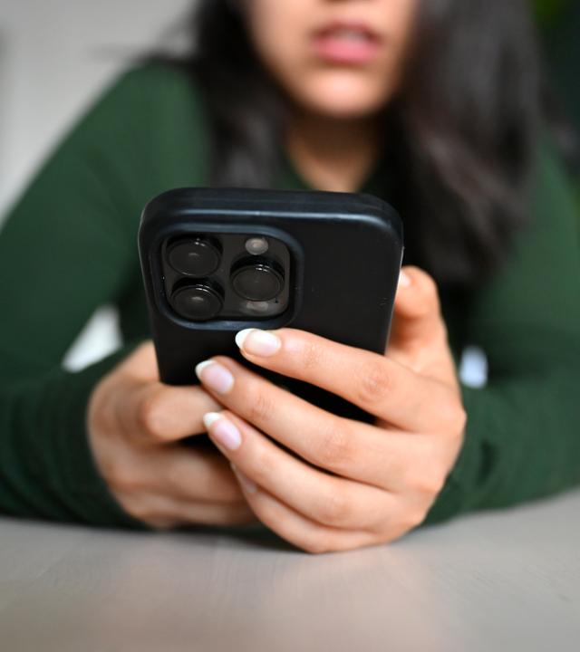 Es ist eine junge Frau zu sehen, wie sie ein schwarzes Handy vor sich in der Hand hält.