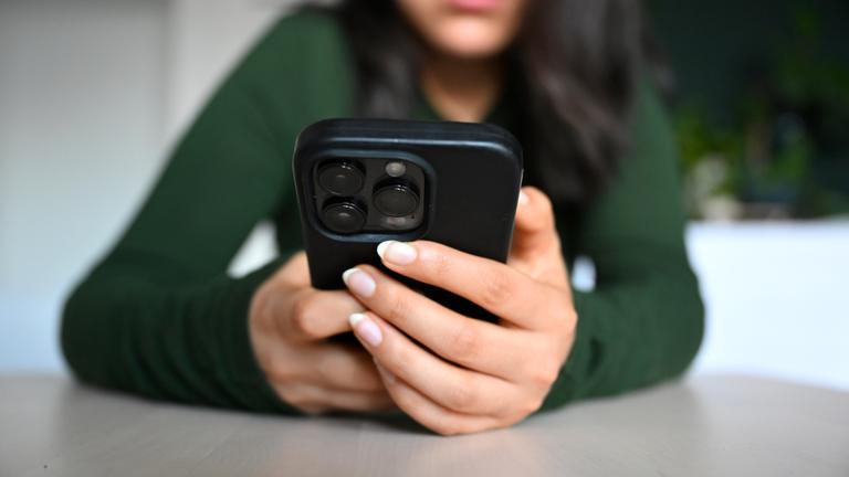 Es ist eine junge Frau zu sehen, wie sie ein schwarzes Handy vor sich in der Hand hält.