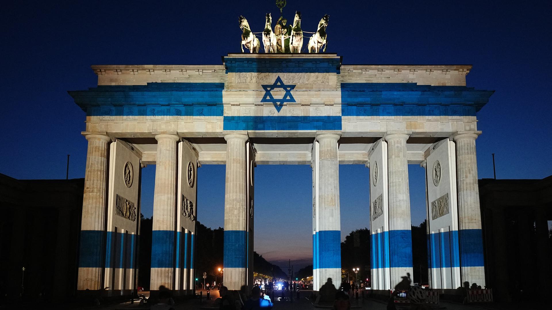 07.10.2026, Berlin: Teilnehmer stehen vor dem mit der Fahne Israels angestrahlten Brandenburger Tor und gedenken bei der Veranstaltung zum Jahrestag des Terrorangriffs der Hamas auf Israel am 7. Oktober 2023 den Opfern.