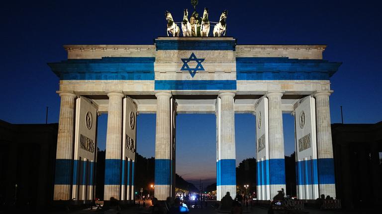 07.10.2026, Berlin: Teilnehmer stehen vor dem mit der Fahne Israels angestrahlten Brandenburger Tor und gedenken bei der Veranstaltung zum Jahrestag des Terrorangriffs der Hamas auf Israel am 7. Oktober 2023 den Opfern.