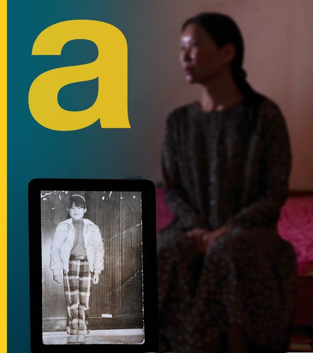 Eine Frau sitzt in einem abgedunkelten Raum, vorne steht ein Tablet und zeigt ein historisches Foto von ihr als Kind.