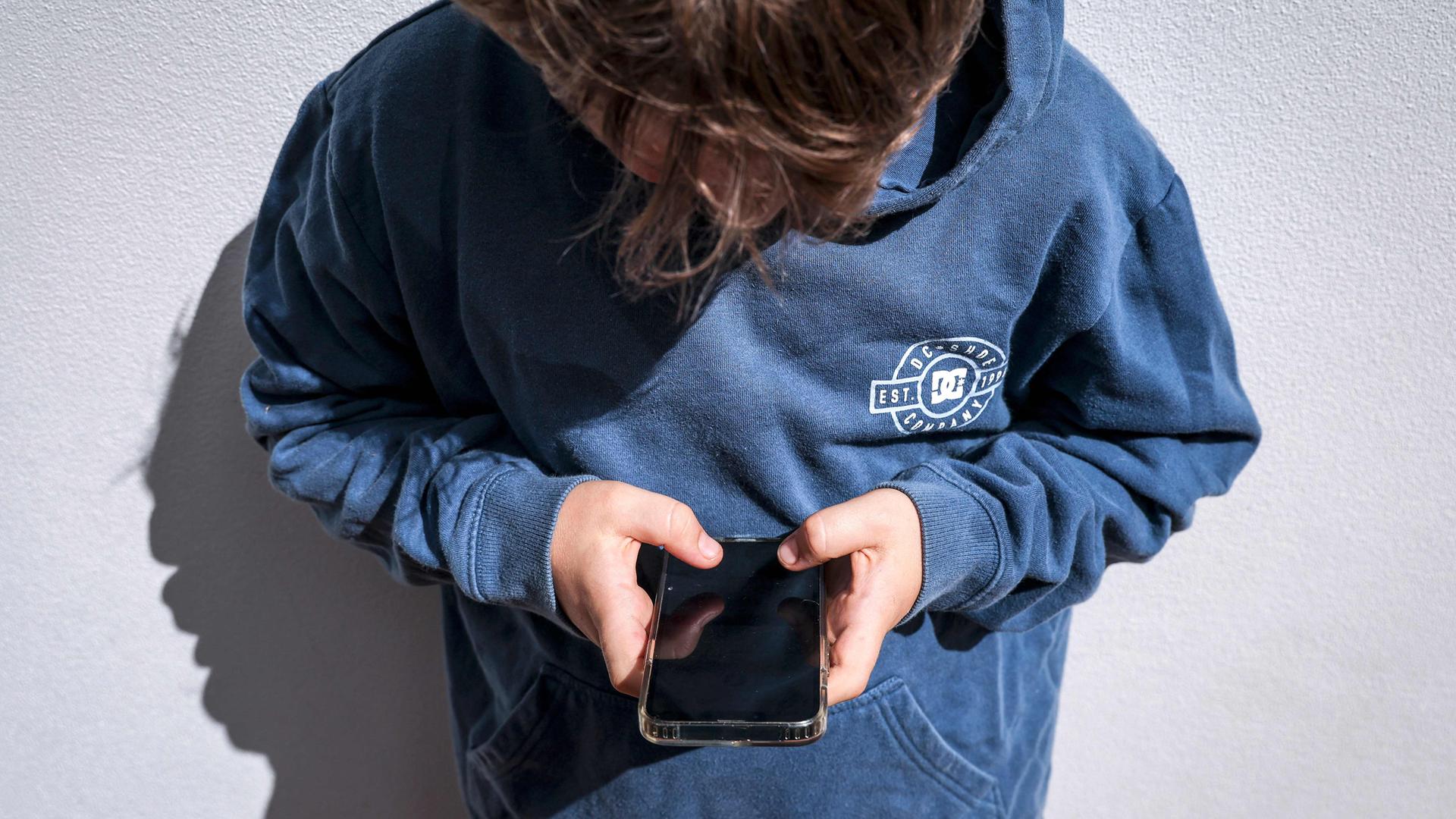 Suchtgefahr bei Jugendlichen: Politik fordert Altersbeschränkungen für Social Media, doch Experten warnen vor einem zu schnellen Verbot. (Bild zeigt eine nach unten aufs Handy schauenden Jungen im blauen Hoodie)