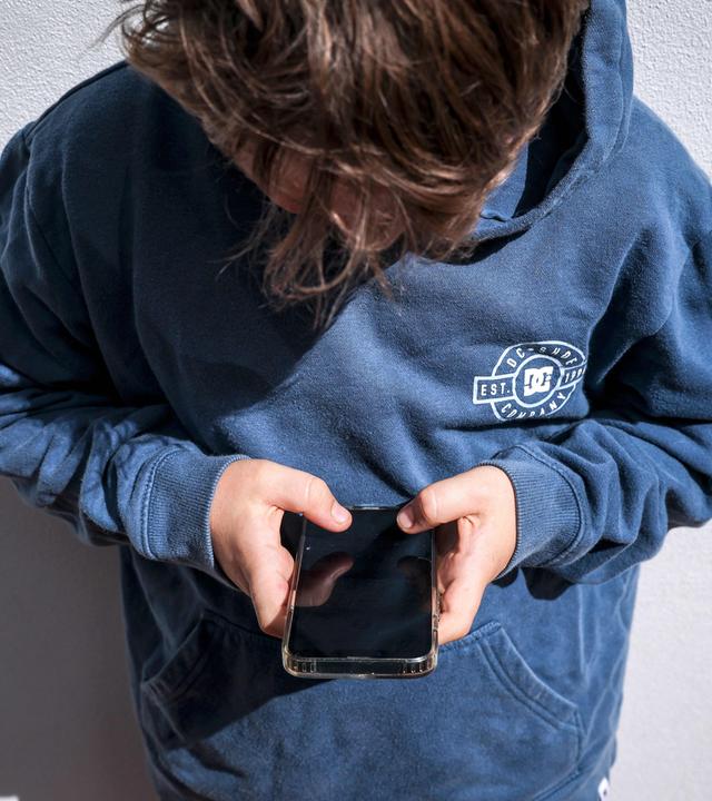 Suchtgefahr bei Jugendlichen: Politik fordert Altersbeschränkungen für Social Media, doch Experten warnen vor einem zu schnellen Verbot. (Bild zeigt eine nach unten aufs Handy schauenden Jungen im blauen Hoodie)