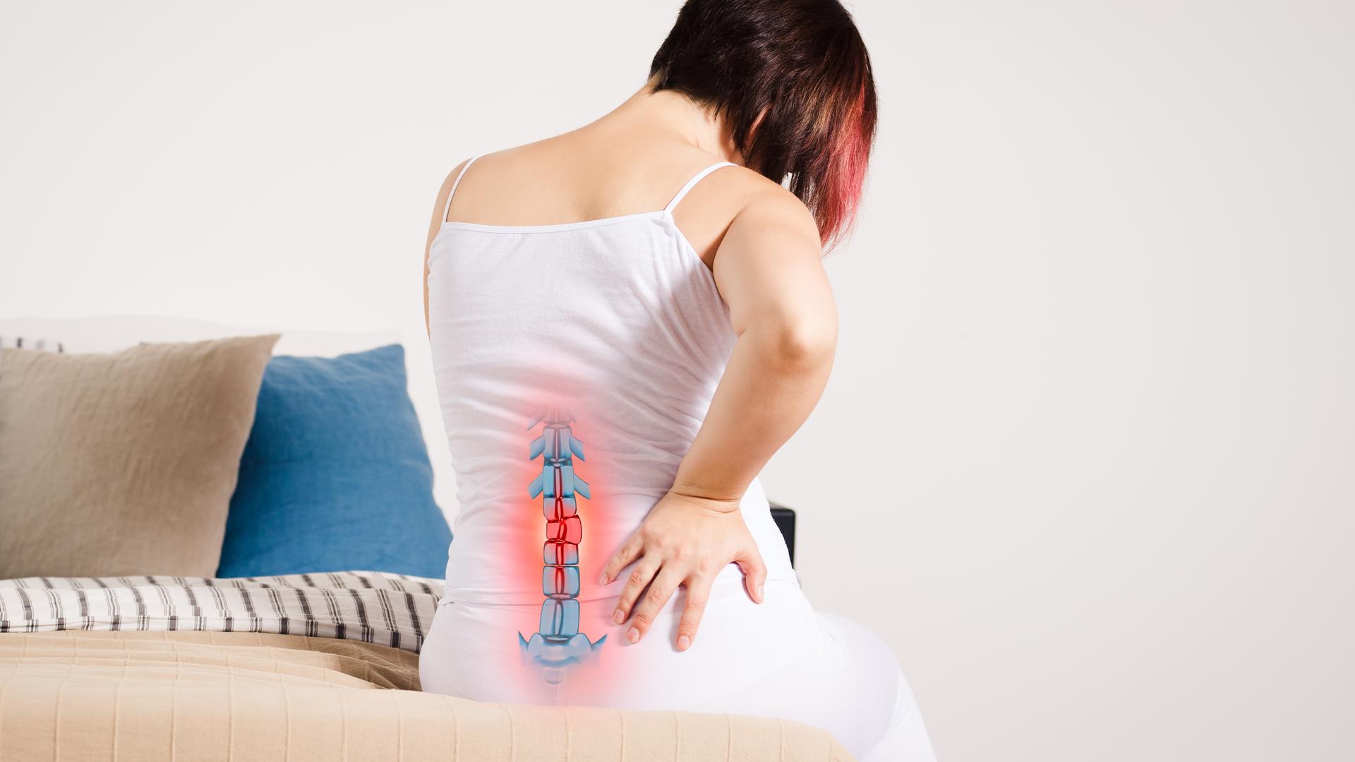 Rückenschmerzen oder Bandscheibenvorfall: Notfall oder reicht Bewegung und Therapie? (Bild: Frau sitzt auf dem Bett, hält den Rücken vor Schmerz - mit hervorgehobenem Skelett)