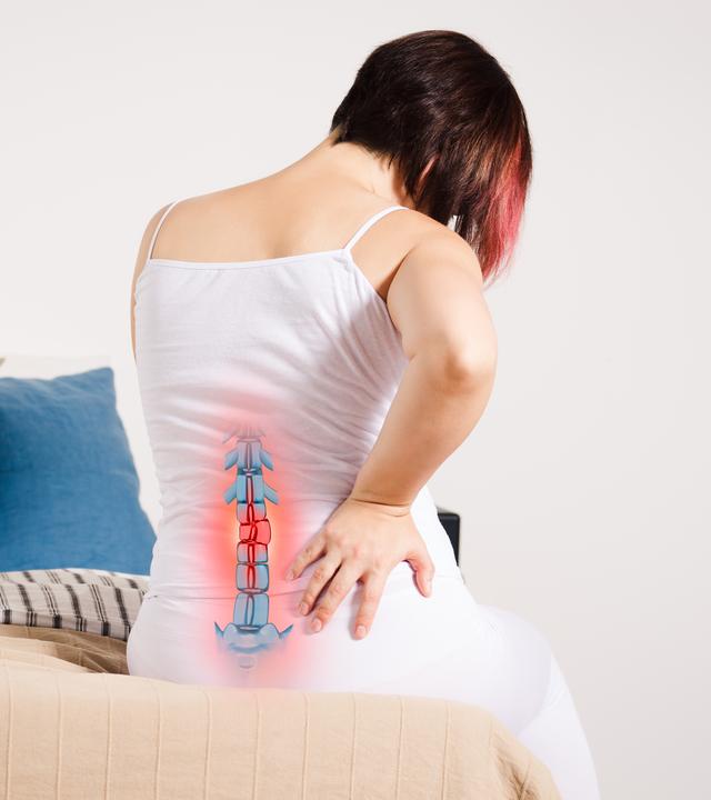 Rückenschmerzen oder Bandscheibenvorfall: Notfall oder reicht Bewegung und Therapie? (Bild: Frau sitzt auf dem Bett, hält den Rücken vor Schmerz - mit hervorgehobenem Skelett)