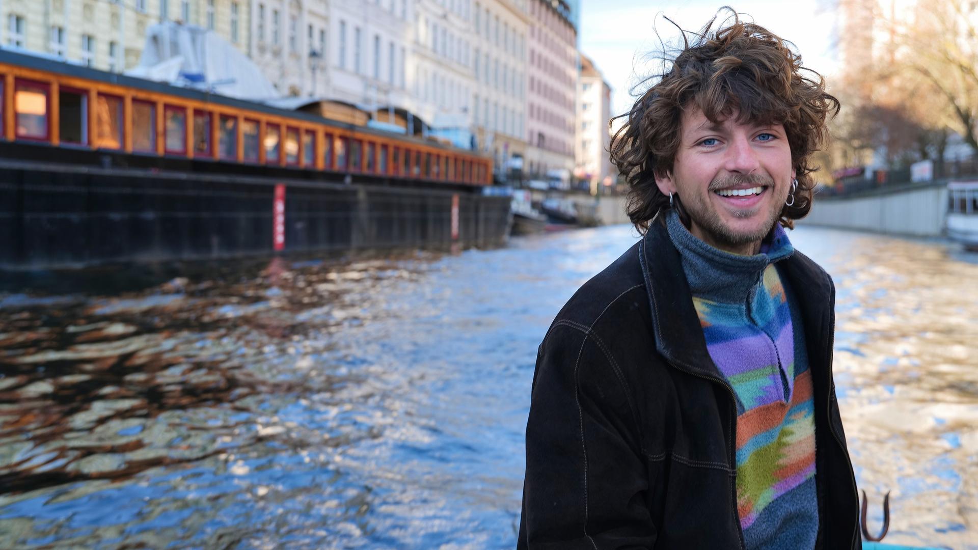 "37°Leben: Die Kraft des Neuanfangs": Linus fährt in einem Boot auf dem Fluss und schaut lachend in die Kamera. An der Seite stehen mehrstöckige Häuser am Fluss, am anderen Ufer einige Bäume.