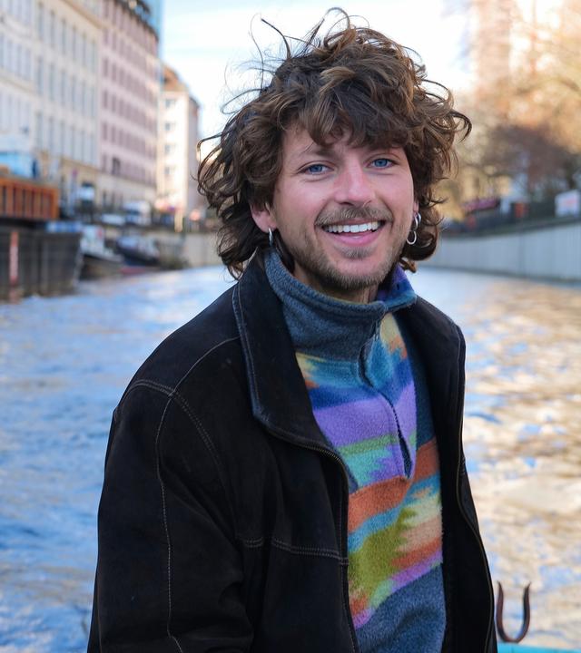 "37°Leben: Die Kraft des Neuanfangs": Linus fährt in einem Boot auf dem Fluss und schaut lachend in die Kamera. An der Seite stehen mehrstöckige Häuser am Fluss, am anderen Ufer einige Bäume.