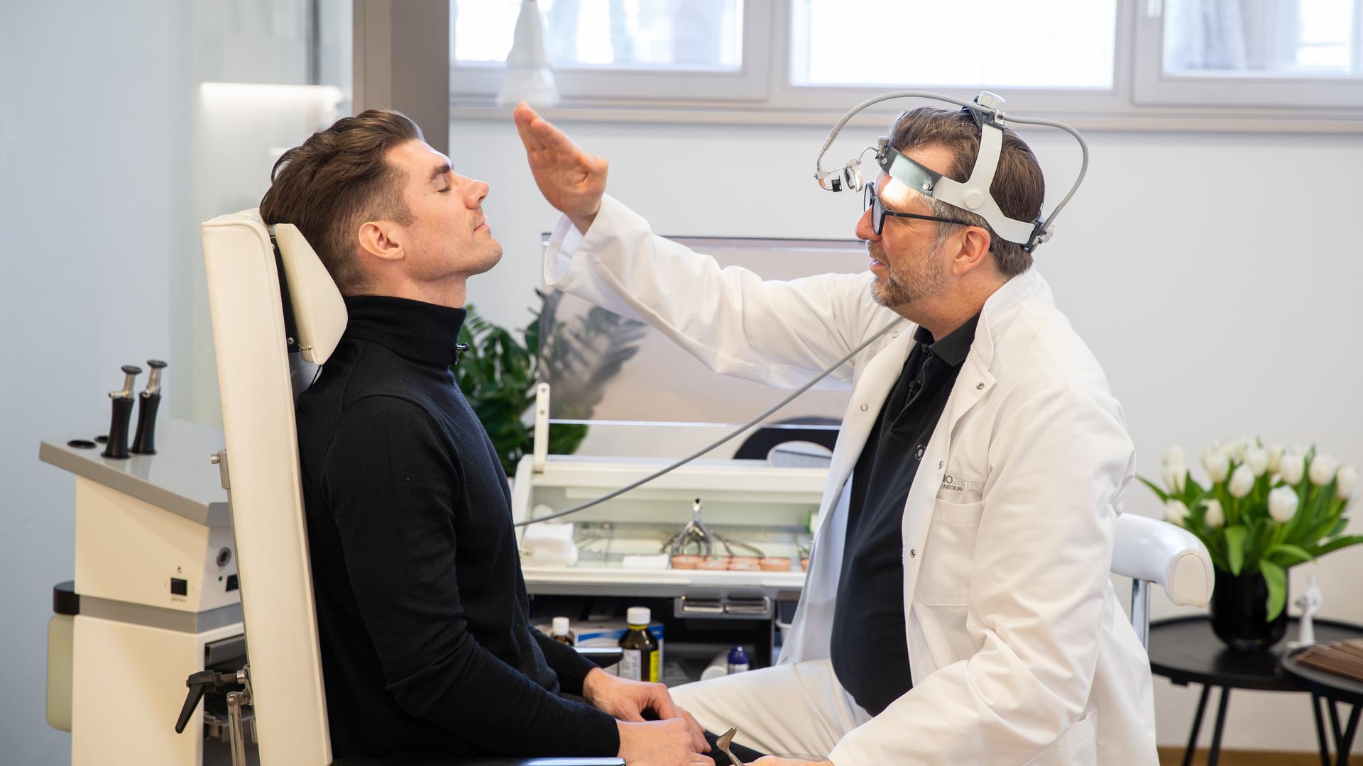 Patient Paul Gressenberger sitzt mit geschlossenen Augen auf einem Behandlungsstuhl. Professor Riedel sitzt vor ihm und schaut sich seine Nase an.