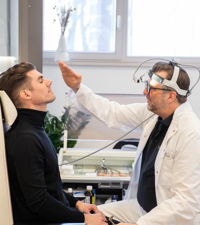 Patient Paul Gressenberger sitzt mit geschlossenen Augen auf einem Behandlungsstuhl. Professor Riedel sitzt vor ihm und schaut sich seine Nase an.