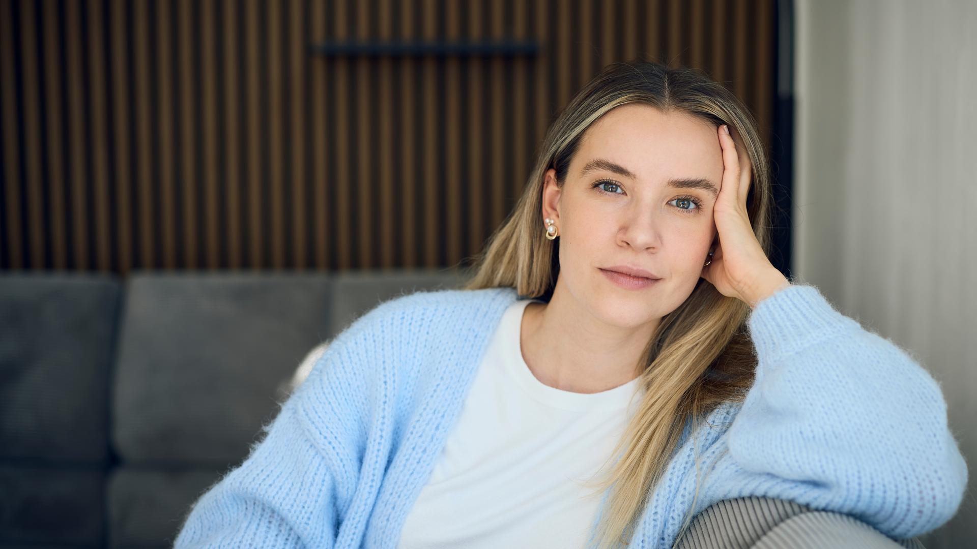 37°Leben: Deutschland, wann bin ich genug?": Valentina, eine blonde junge Frau sitzt auf einem grauen Sofa in einer modernen Wohnung. Sie stützt ihren Kopf mit der Hand ab und blickt ruhig und nachdenklich in die Kamera.