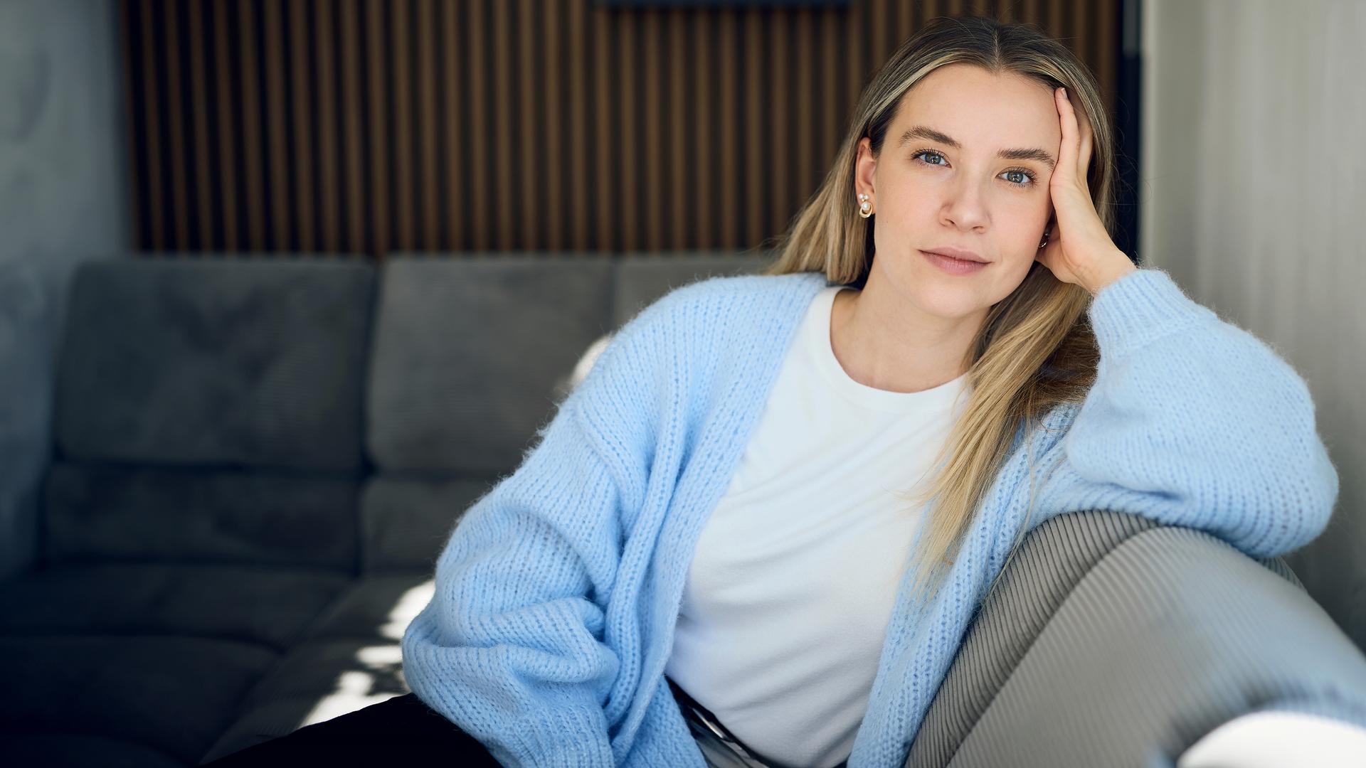 37°Leben: Deutschland, wann bin ich genug?": Valentina, eine blonde junge Frau sitzt auf einem grauen Sofa in einer modernen Wohnung. Sie stützt ihren Kopf mit der Hand ab und blickt ruhig und nachdenklich in die Kamera.