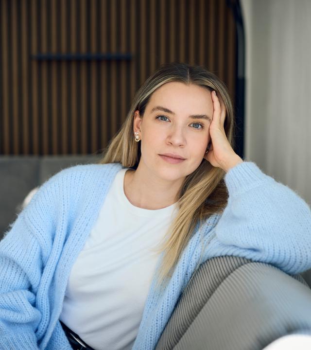 37°Leben: Deutschland, wann bin ich genug?": Valentina, eine blonde junge Frau sitzt auf einem grauen Sofa in einer modernen Wohnung. Sie stützt ihren Kopf mit der Hand ab und blickt ruhig und nachdenklich in die Kamera.